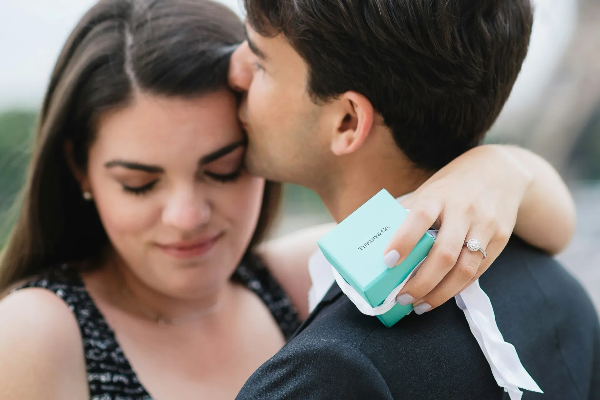 Couple embracing with woman holding a Tiffany blue box and wearing a diamond engagement ring