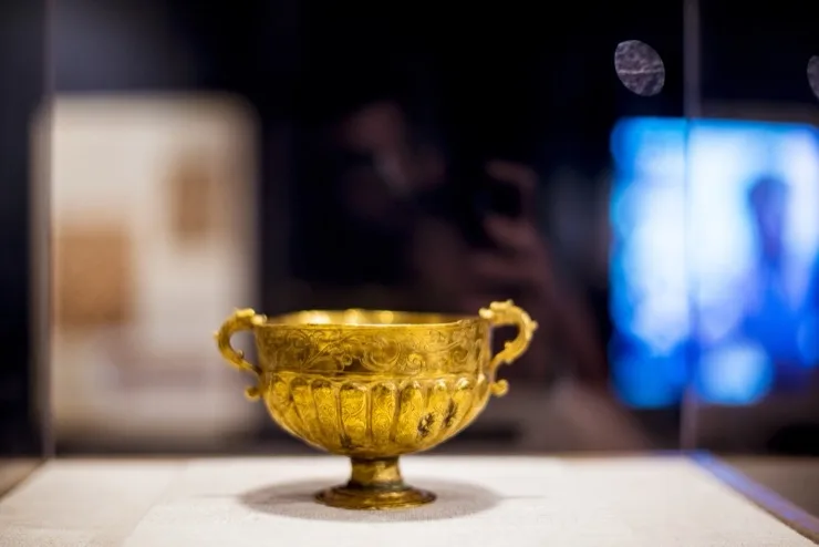 Ornate gold chalice with floral repousse work and handles displayed in a museum case