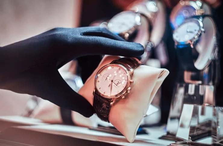 Gloved hand presenting a rose gold luxury chronograph watch on a white display cushion